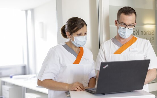 Zwei medizinische Fachkräfte in weiß-orangener Klinikbekleidung mit Masken dokumentieren Patientendaten am Laptop im Krankenhaus