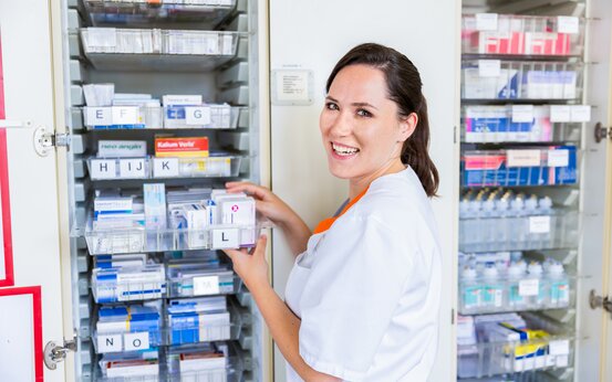 Pflegekraft in weißer Kleidung sortiert Medikamente in offenem Medikamentenschrank mit beschrifteten Fächern
