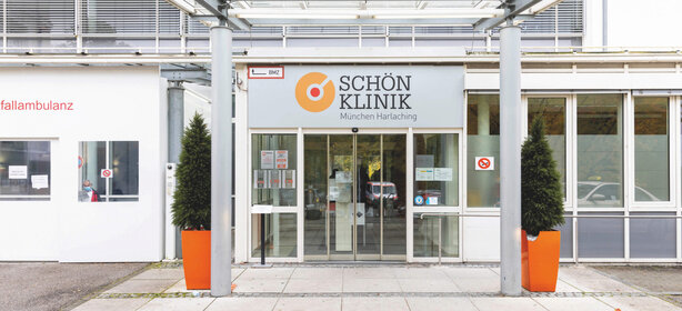 Haupteingang der Schön Klinik München Harlaching mit Glasdach, zwei großen Pflanzen in Pflanzkübeln, Glastür und Klinik-Logo über dem Eingang