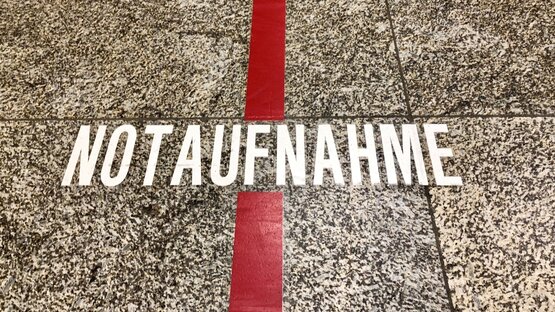 Schriftzug „Notaufnahme“ in weißen Großbuchstaben auf grau-gesprenkeltem Boden, breite rote Linie verläuft senkrecht durch das Wort