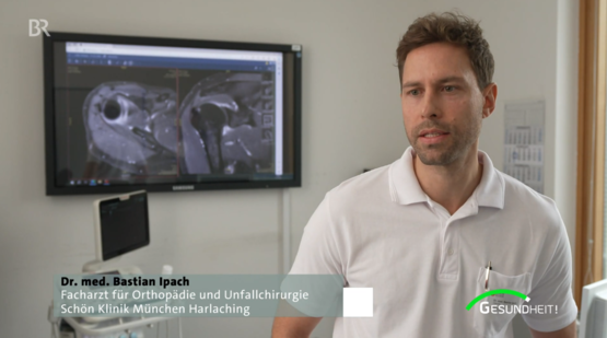 Dr. Bastian Ipach spricht in moderner Praxis der Schön Klinik München Harlaching, Monitor mit MRT-Bildern im Hintergrund