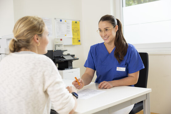 Patientin und Anästhesistin im Aufklärungsgespräch an weißem Tisch, Anästhesistin in blauer Kleidung mit Namensschild