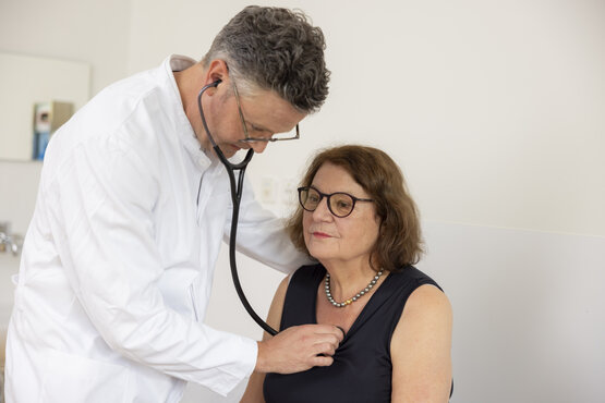 Frau mit Brille sitzt in Klinik, Arzt im weißen Kittel hört mit Stethoskop ihre Brust ab, medizinisches Untersuchungsgespräch