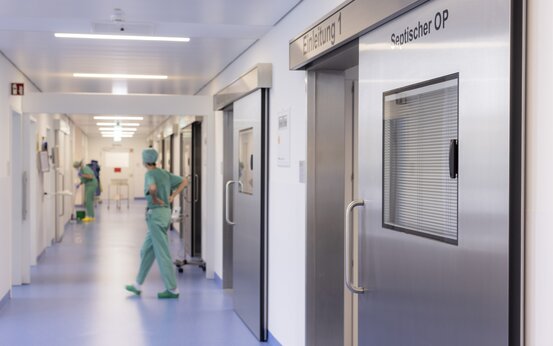 Krankenhausflur mit blauem Boden, weißen Wänden, Edelstahltüren „Einleitung 1“ und „Septischer OP“, dahinter OP-Personal in grüner Kleidung