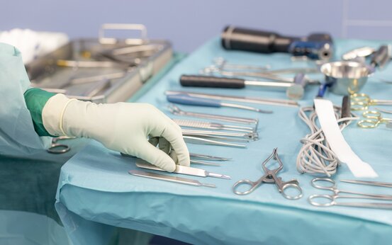 Hand mit OP-Handschuh greift auf OP-Tisch nach Skalpell, umgeben von chirurgischen Instrumenten auf blauem sterilen Tuch