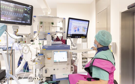 Medizinisches Fachpersonal mit grünem Haarnetz, OP-Maske und pinkfarbener Schutzkleidung sitzt an Arbeitsplatz mit Monitoren und Geräten im Klinikraum