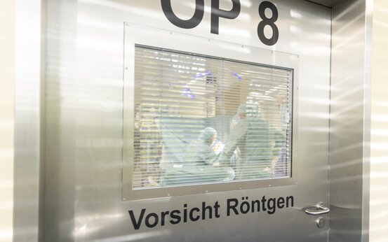 Edelstahl-Tür mit der Aufschrift „OP 8“ und „Vorsicht Röntgen“, Fenster mit Jalousie, dahinter mehrere Personen in grüner OP-Kleidung