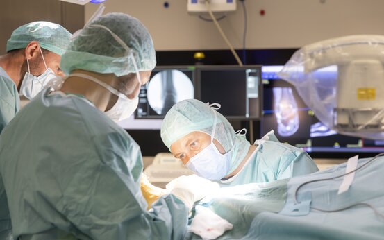 Drei Chirurgen in OP-Kleidung und Mundschutz führen gemeinsam eine orthopädische Operation durch, im Hintergrund Monitore mit Röntgenbildern 