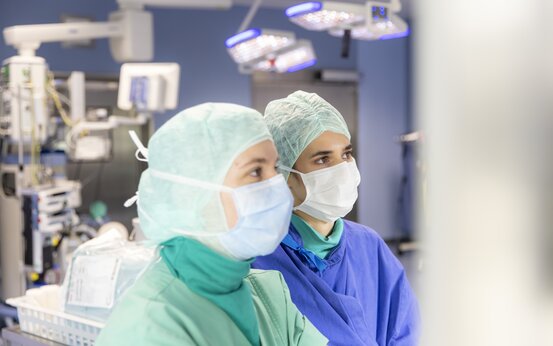 Zwei medizinische Fachkräfte in OP-Kleidung mit Mund-Nasen-Schutz und Haube stehen im modernen Operationssaal, umgeben von Geräten und Monitoren