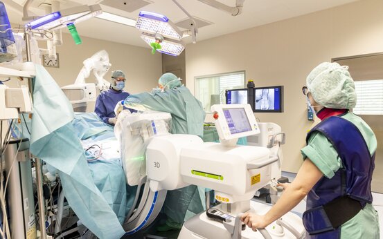 Drei Personen in OP-Kleidung und Masken operieren im modernen Operationssaal mit Röntgengerät und Monitoren