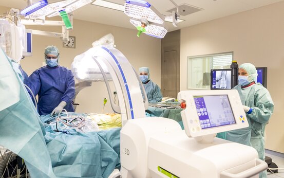 Drei Personen in OP-Kleidung stehen im hellen Operationssaal um einen Patienten mit blauen Tüchern