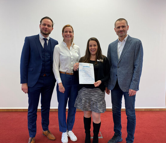Vier Personen auf rotem Teppich, zwei Männer im Anzug, zwei Frauen in Business-Kleidung, mittig Zertifikat