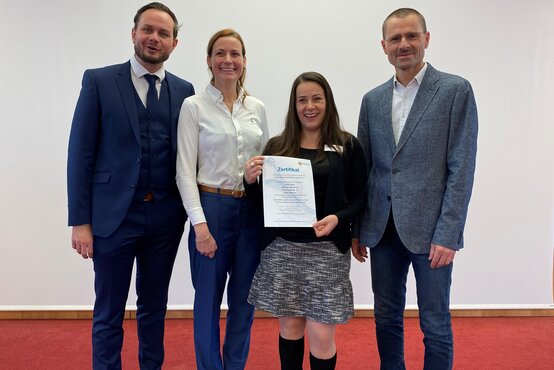 Vier Personen auf rotem Teppich, zwei Männer im Anzug, zwei Frauen in Business-Kleidung, mittig Zertifikat