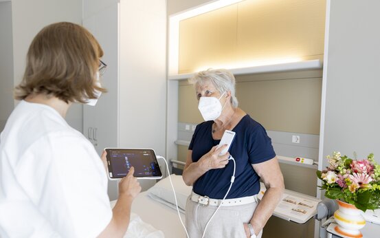 Ärztin und Patientin mit Mundschutz im Krankenhauszimmer, Patientin nutzt medizinisches Gerät am Oberkörper