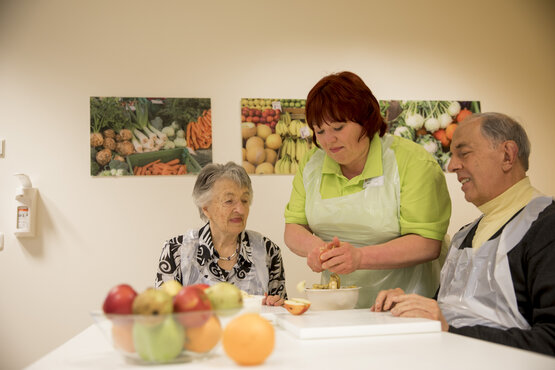 Pflegerin hilft älteren Menschen beim Apfelschneiden am Tisch in der Demenz-Tagesklinik, Obstschale und bunte Bilder im Hintergrund