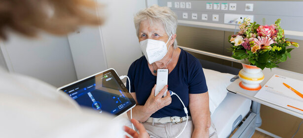Ältere Frau mit Maske sitzt im Krankenhausbett und hält medizinisches Gerät am Hals