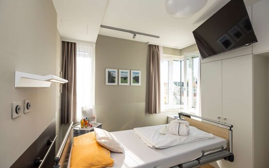 Modernes Patientenzimmer mit Pflegebett, gelbem Kissen, Tageslicht, Schrank und Fernseher