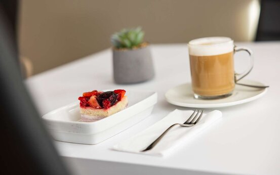 Weiß gedeckter Tisch mit Stück Obstkuchen auf rechteckigem Teller, Cappuccino in Glastasse und Sukkulente im Blumentopf