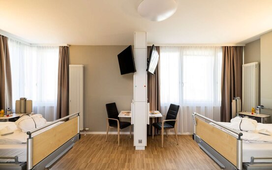 Doppelzimmer mit zwei Pflegebetten, Trennwand mit TV, Esstisch, großen Fenstern und Tageslicht