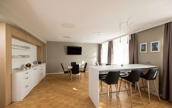 Moderner, heller Loungebereich mit weißer Anrichte, Barhockern, Obstkorb, Tischen, schwarzen Stühlen und großem Fenster