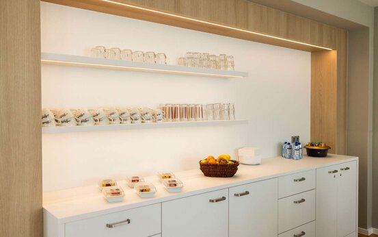 Heller Loungebereich mit weißen Schränken, Holzrückwand, Regalen mit Gläsern und Tassen, Schalen mit Speisen und Obstkorb