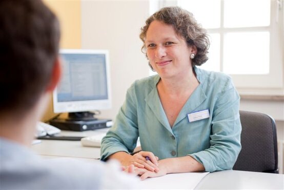 Frau mit lockigen Haaren und Namensschild sitzt an einem Schreibtisch in hellem Büro, lächelt eine Person im Vordergrund an