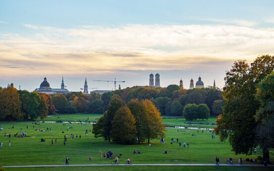 Menschen unterschiedlichen Alters im Englischen Garten München, Skyline mit Frauenkirche im Hintergrund.
