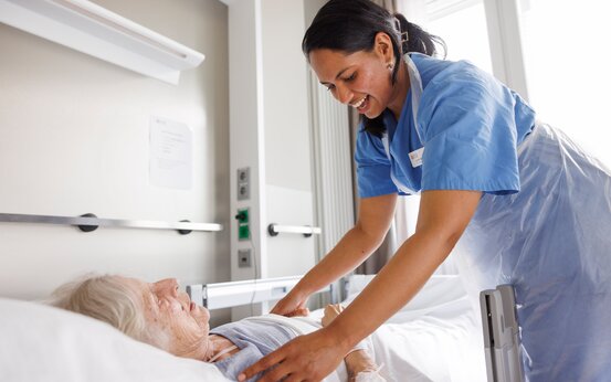 Pflegekraft versorgt bettlägerige Person im hellen Krankenhauszimmer mit Notrufknopf und Tageslicht.