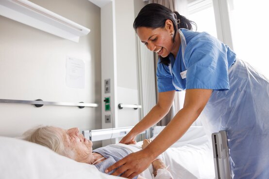 Pflegekraft versorgt bettlägerige Person im hellen Krankenhauszimmer mit Notrufknopf und Tageslicht.