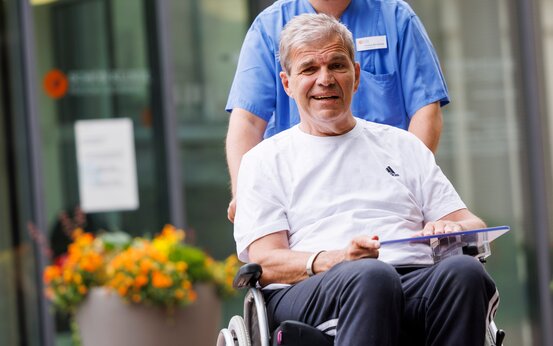  Pflegekraft schiebt Patient im Rollstuhl vor den Eingang der SCHÖN KLINIK München Schwabing.