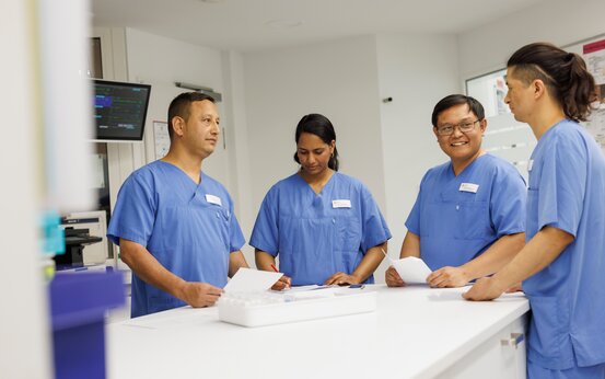 Vier Pflegekräfte im blauen Kasack besprechen Unterlagen am Tresen in moderner Klinik, Teamarbeit im Pflegealltag.