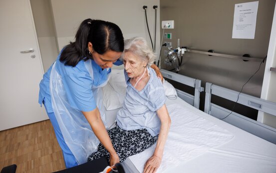 Pflegekraft hilft älterer Patientin beim Aufrichten im Klinikbett, individuelle Unterstützung in der Frührehabilitation.