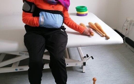Frau mit Armschlinge trainiert auf Therapieliege mit Holzkeulen und bunten Ringen in der Frührehabilitation.