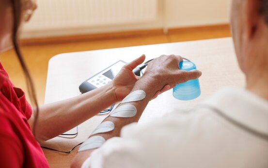 Elektrostimulation am Unterarm eines Patienten mit Greifübung eines blauen Bechers unter therapeutischer Anleitung.