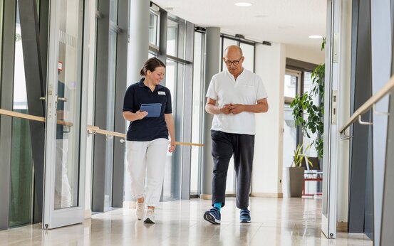 Therapeutin begleitet Parkinson-Patient im modernen Klinikflur mit Handläufen und Tageslicht.