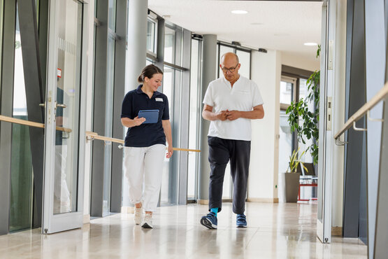 Therapeutin begleitet Parkinson-Patient im modernen Klinikflur mit Handläufen und Tageslicht.