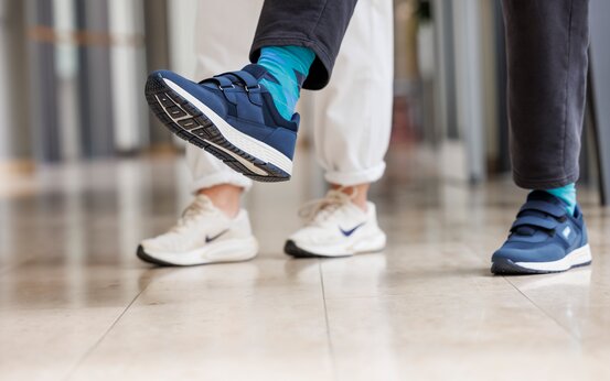 Zwei Personen gehen im Klinikflur, Fokus auf Antifreezingschuh und Nike-Sneaker.