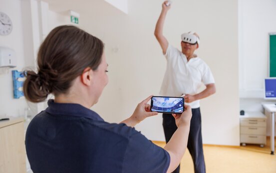 Person mit VR-Brille und Ball macht Bewegungstraining, zweite Person überwacht per Smartphone.