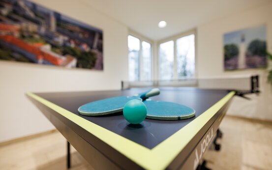 Zwei grüne Tischtennisschläger und Ball auf Tischtennisplatte in hellem Raum, Bewegungsförderung bei Parkinson.