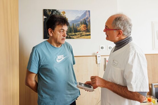 Patient mit Parkinson im Gespräch, Mediziner demonstriert Steuergerät für Therapieeinstellung im Behandlungsraum.