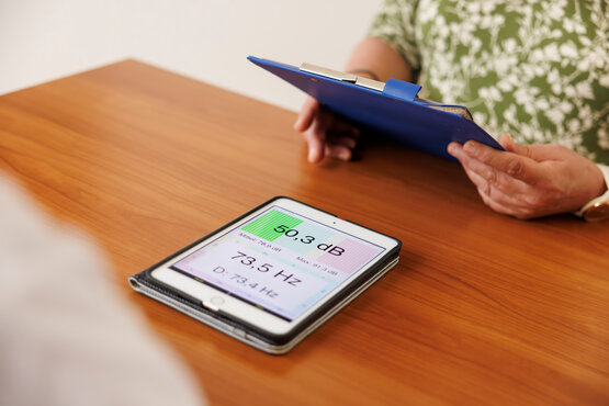 Tablet mit Lautstärke- und Frequenzanalyse, Logopädin mit Klemmbrett erklärt Patientin Therapie bei Parkinson in Klinik.