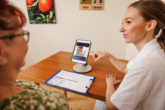 Logopädin und Patientin analysieren Gangmuster und Sprachübungen bei Parkinson, Tablet und Therapiemappe auf Tisch in Klinik.
