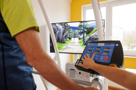 Patient mit Parkinson trainiert das Gehen auf modernem Gangtrainer mit virtueller Landschaft, betreut von Therapeutin in heller Klinik.