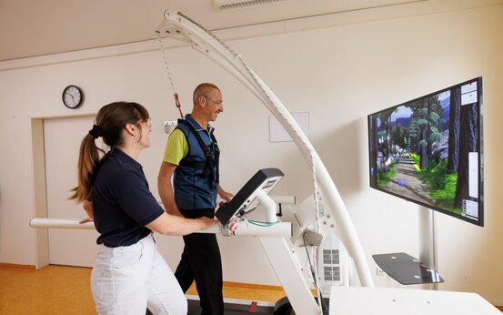 Patient mit Parkinson auf Laufband, gesichertem Gangtrainer und virtuellem Landschaftsbild, betreut von Therapeutin in Klinik.