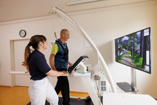 Patient mit Parkinson auf Laufband, gesichertem Gangtrainer und virtuellem Landschaftsbild, betreut von Therapeutin in Klinik.