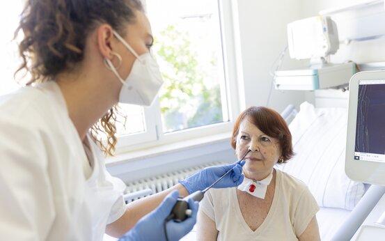 Medizinische Fachkraft untersucht ältere Frau mit Trachealkanüle per Nasensonde im Krankenbett, medizinische Geräte sichtbar