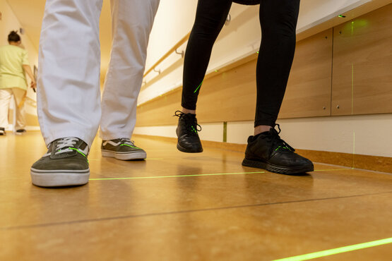 Zwei Personen gehen im Klinikflur über grüne Laserlinien zur Gangschulung, Handlauf an der Wand