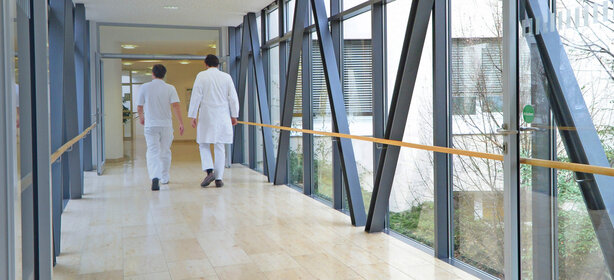 Zwei Personen in weißer Kleidung gehen durch hellen Klinikflur mit bodentiefen Fenstern und Handläufen