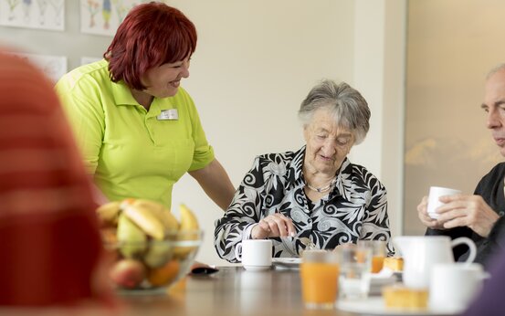 Pflegekraft und älterer Frau beim Kaffeetrinken im Gemeinschaftsraum, gedeckter Tisch mit Kuchen und Obst