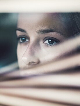 Nahaufnahme eines Gesichts, das durch geöffnete Jalousien blickt, Hand schiebt Lamellen auseinander, Gesicht teilweise verdeckt